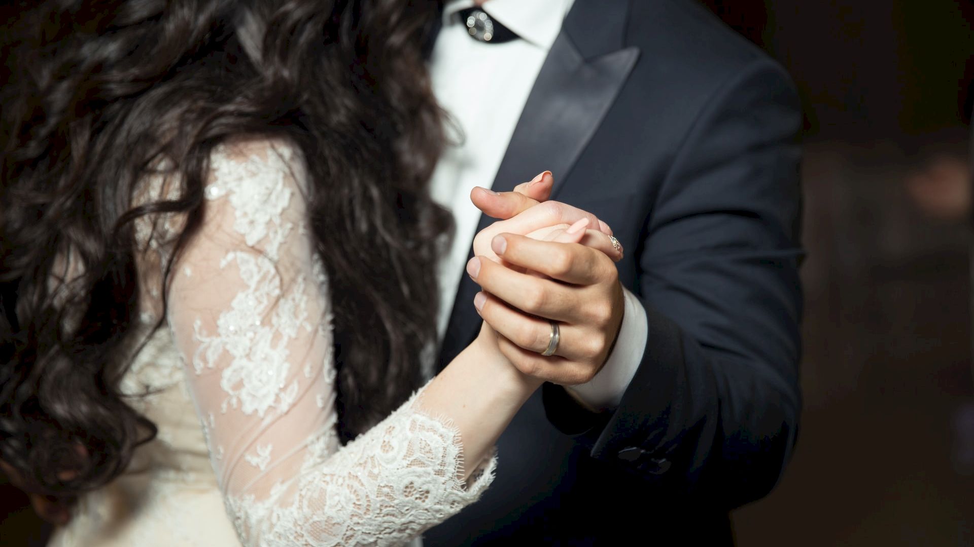 A couple is dancing closely, with the woman in a white lace dress and the man in a dark suit, holding hands lovingly.