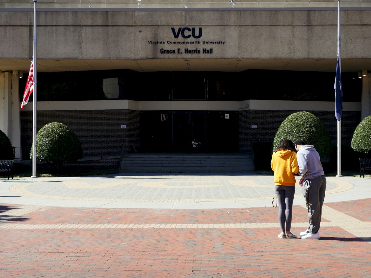 The image shows two people standing in front of Virginia Commonwealth University's Grace E. Harris Hall. They are facing the building.