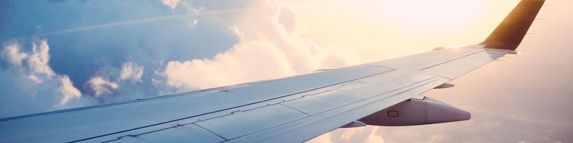 The image shows the wing of an airplane flying above the clouds with a beautiful sunrise or sunset in the background, illuminating the sky and clouds.