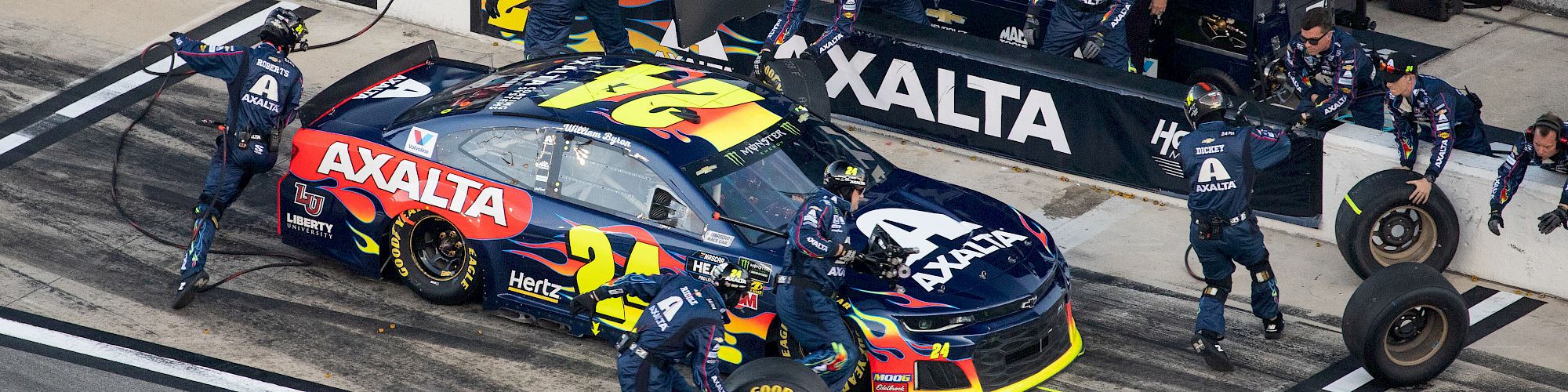 A race car is in the pit stop, surrounded by the crew changing tires and refueling. The car and pit box have prominent "AXALTA" branding.