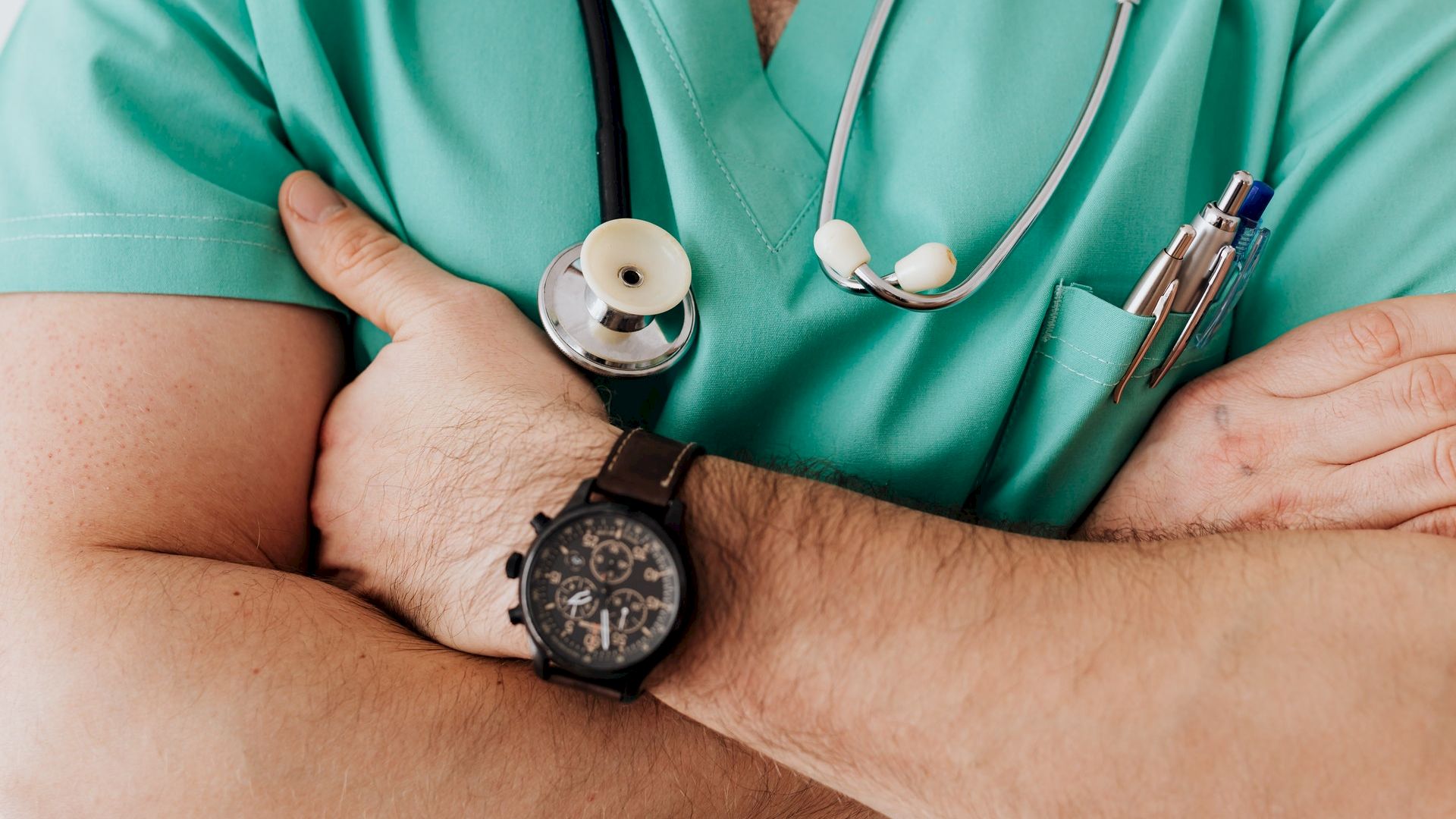 The image shows a person in green scrubs with a stethoscope around their neck, arms crossed, and a watch on the wrist, holding some pens.