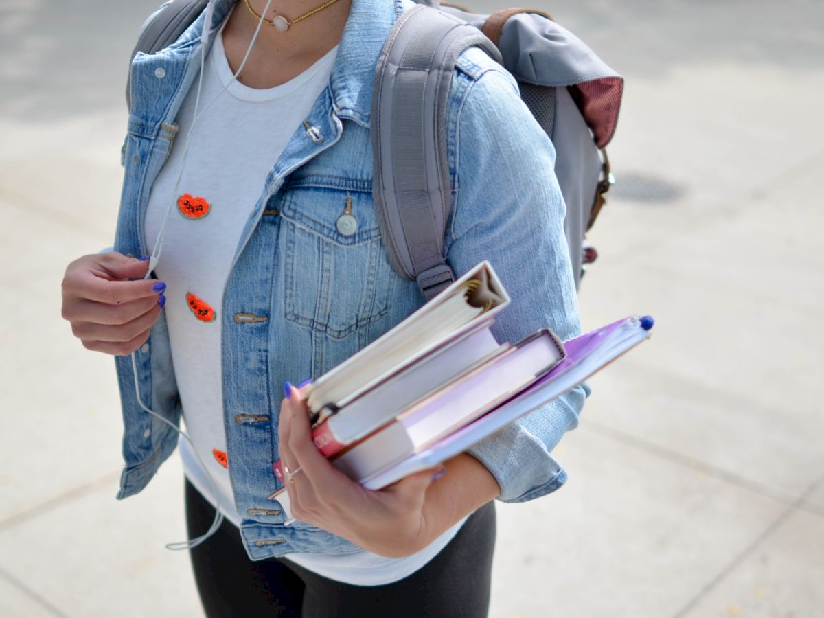 A person wearing a denim jacket and a backpack holds several books in one arm, with a light background and surface.