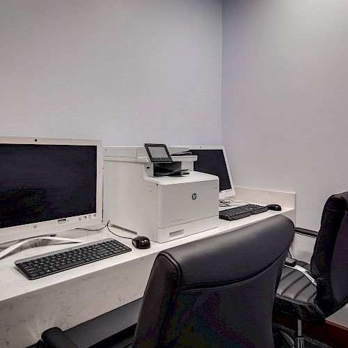 This image shows a small office setup with two computers, a white printer, and two black office chairs against a plain wall.