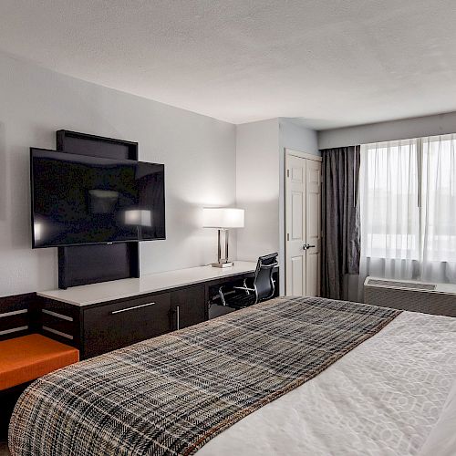 This image shows a modern hotel room with a large bed, wall-mounted TV, desk, chair, bench, artwork, and a window with curtains.