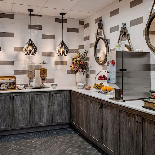 A well-organized breakfast buffet area with cereal dispensers, fruits, a juice machine, and elegant lighting and decor, including mirrors and flowers.