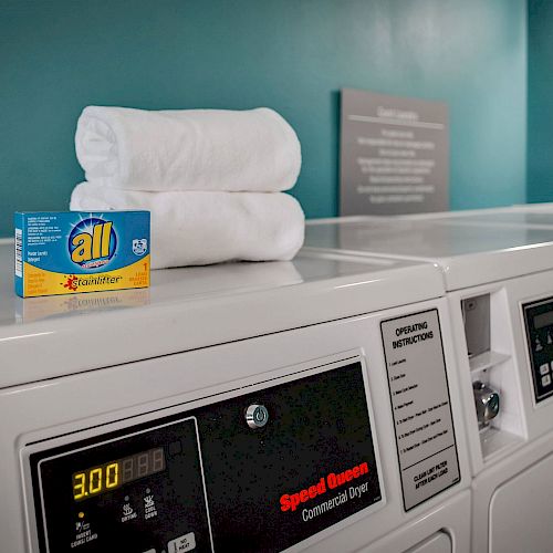 The image shows a laundry room with washing machines, a box of detergent, and two folded white towels on top of one of the machines.