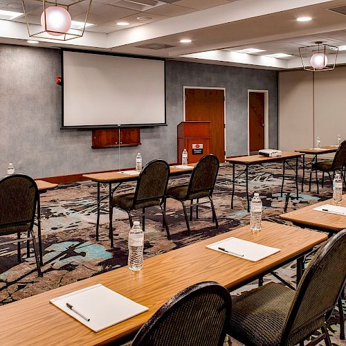 A conference room setup with rows of tables and chairs, a projector screen at the front, and water bottles and notepads placed on the tables.