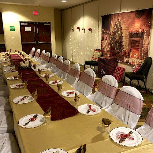 A long table is set with white plates, red and gold decorations, and chairs dressed in white and gold covers, ready for a festive dinner event.