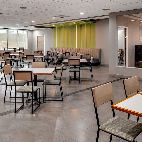 The image shows a large, empty cafeteria with several tables and chairs, modern decor, large windows, and a section with booth seating.