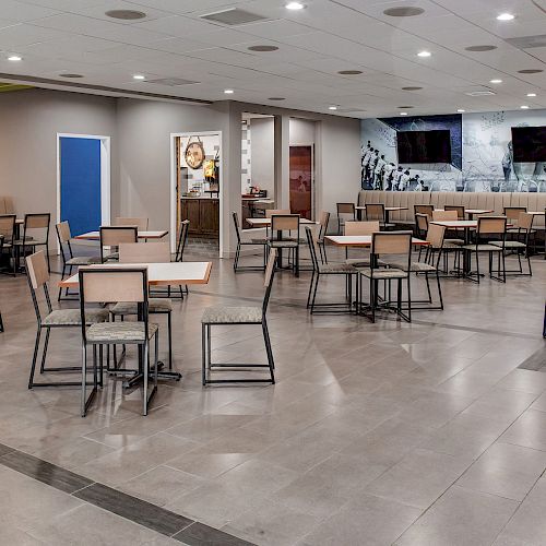 The image shows an empty modern dining area with tables and chairs, a bar with stools, and a wall with two flat-screen televisions.