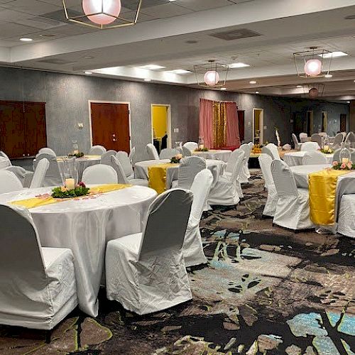 A well-decorated banquet hall with round tables covered in white tablecloths, chairs with white covers, and yellow table runners and floral centerpieces.