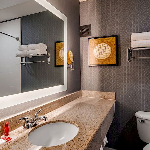 The image shows a bathroom with a sink, mirror, toiletries, towel rack with towels, a toilet, and wall decor.