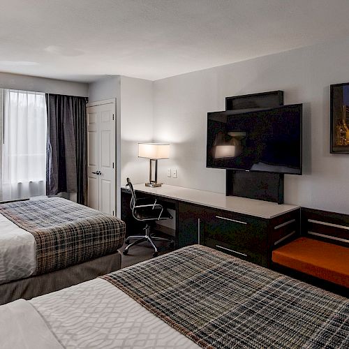 The image shows a hotel room with two beds, a TV, a desk with a chair, a small sofa, and a lamp by the window.