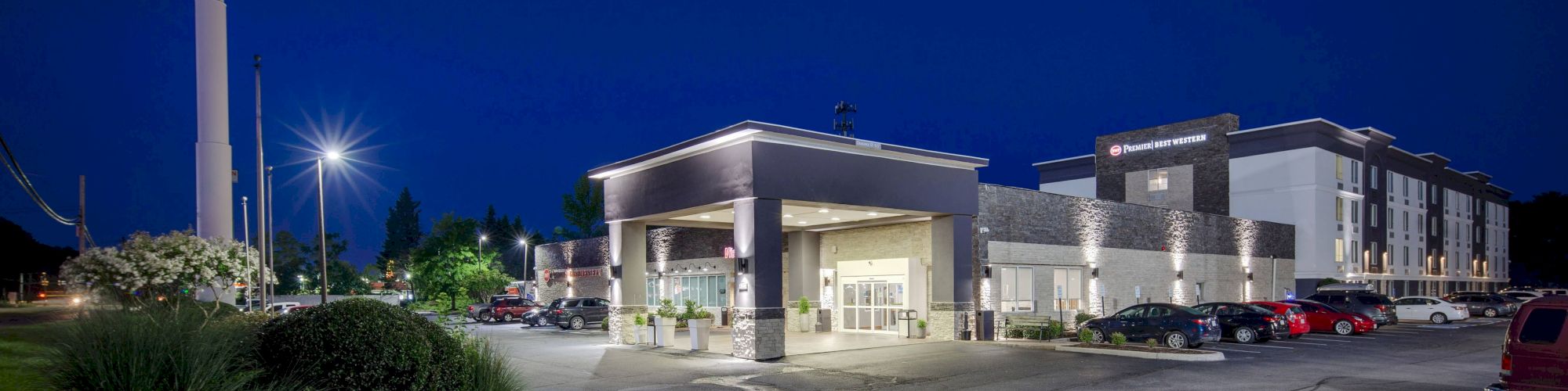 The image shows a well-lit hotel building with a driveway and parked cars in front, set against a dark blue evening sky.