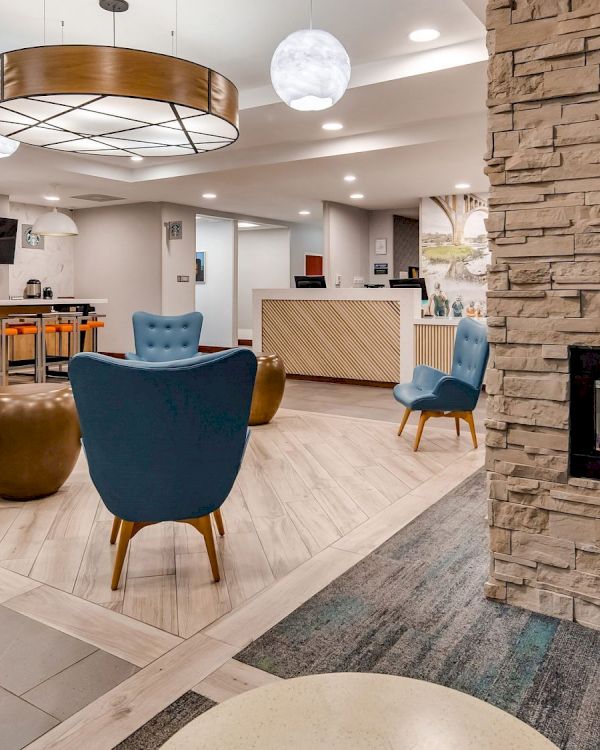 A modern lounge area features blue chairs and a stone fireplace, with a reception desk and a bar counter in the background.