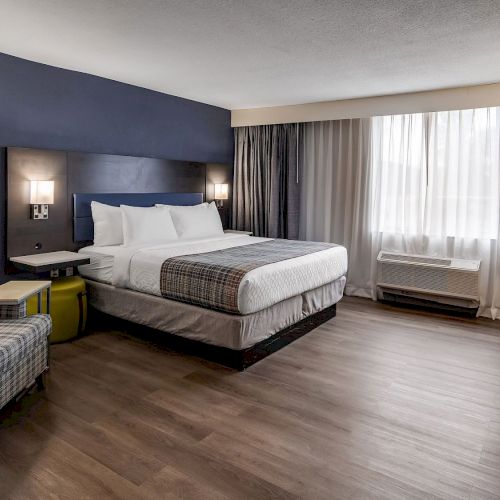 A modern hotel room with a large bed, a plaid armchair, bedside lamps, a desk, and a window with curtains, featuring wood flooring.