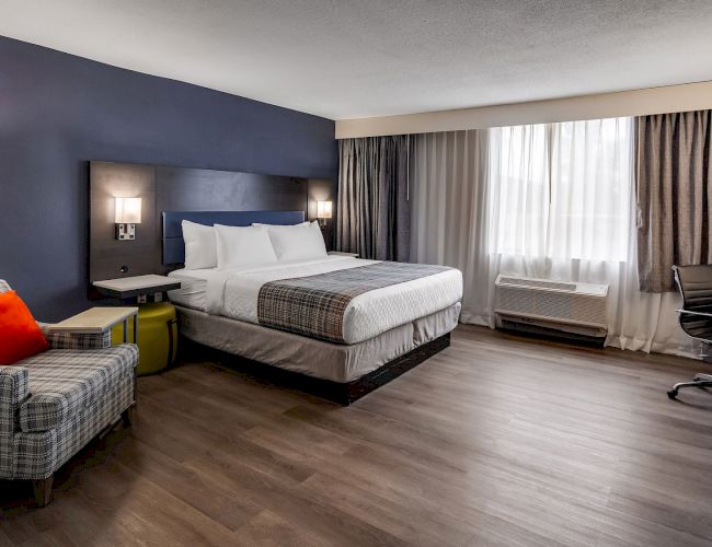 A modern hotel room with a large bed, a plaid armchair, bedside lamps, a desk, and a window with curtains, featuring wood flooring.