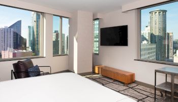 A modern hotel room features large windows with a city view, a bed, a TV mounted on the wall, and minimalistic decor.
