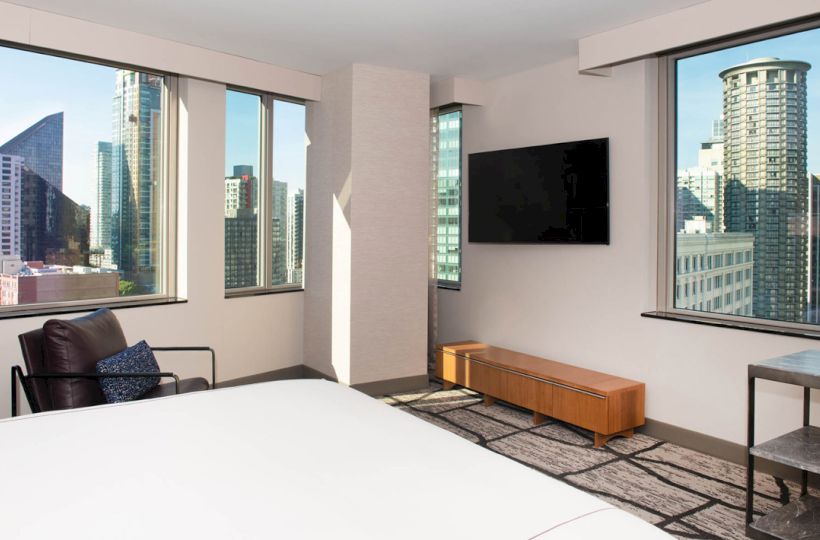 A modern hotel room features large windows with a city view, a bed, a TV mounted on the wall, and minimalistic decor.