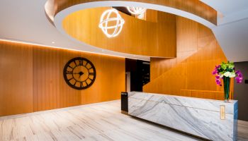 A modern lobby with wooden panels, a spiral staircase, a large wall clock, a marble reception desk, and a vase of flowers.