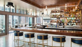 A modern bar with a wooden counter and high stools, various bottles on display behind the bar, and large windows providing a city view ends the sentence.