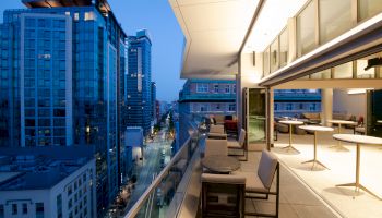 A modern urban apartment balcony overlooking a city street at dusk, with tables, chairs, and view of high-rise buildings illuminated by city lights.