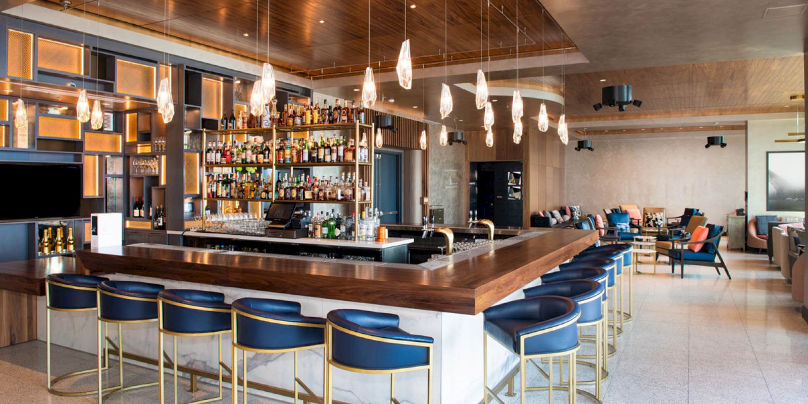 A modern bar with blue cushioned stools, pendant lights, and a stocked bar counter in a spacious area with additional seating.