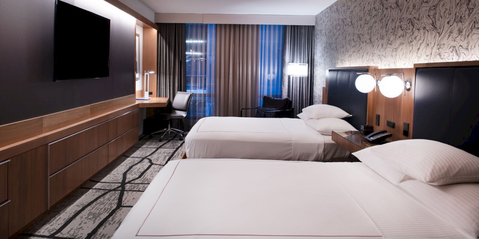 A modern hotel room with two neatly made beds, a flat-screen TV, a desk with a chair near the window, stylish decor, and ambient lighting.