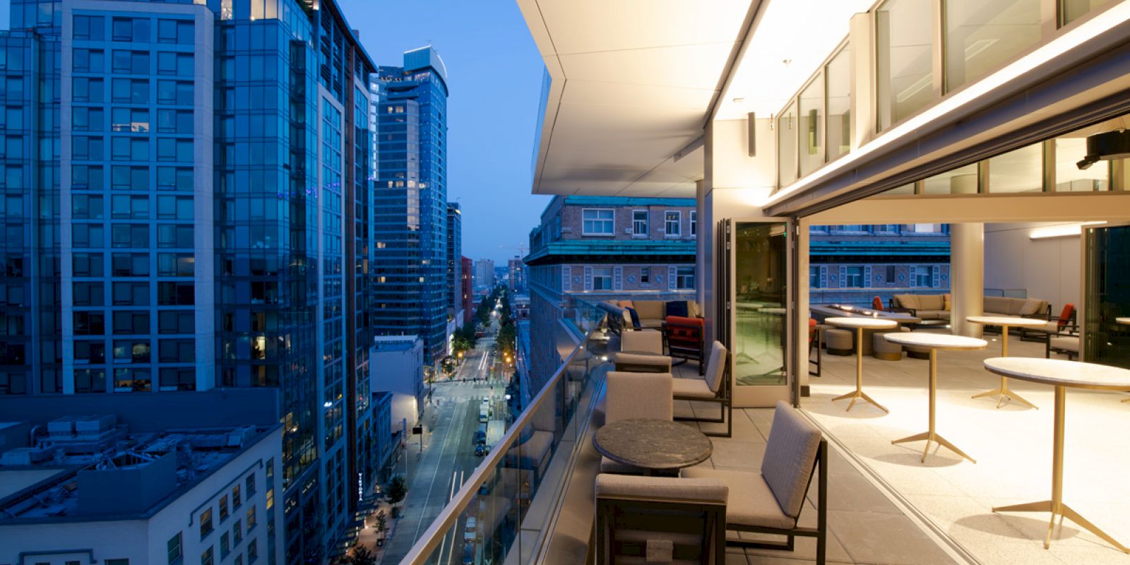 The image shows a modern cityscape at dusk with tall buildings, and a spacious balcony with seating and tables overlooking the street below.