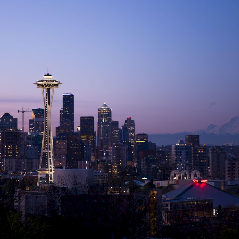 The image shows the skyline of Seattle at twilight, featuring the Space Needle prominently with Mount Rainier visible in the background.