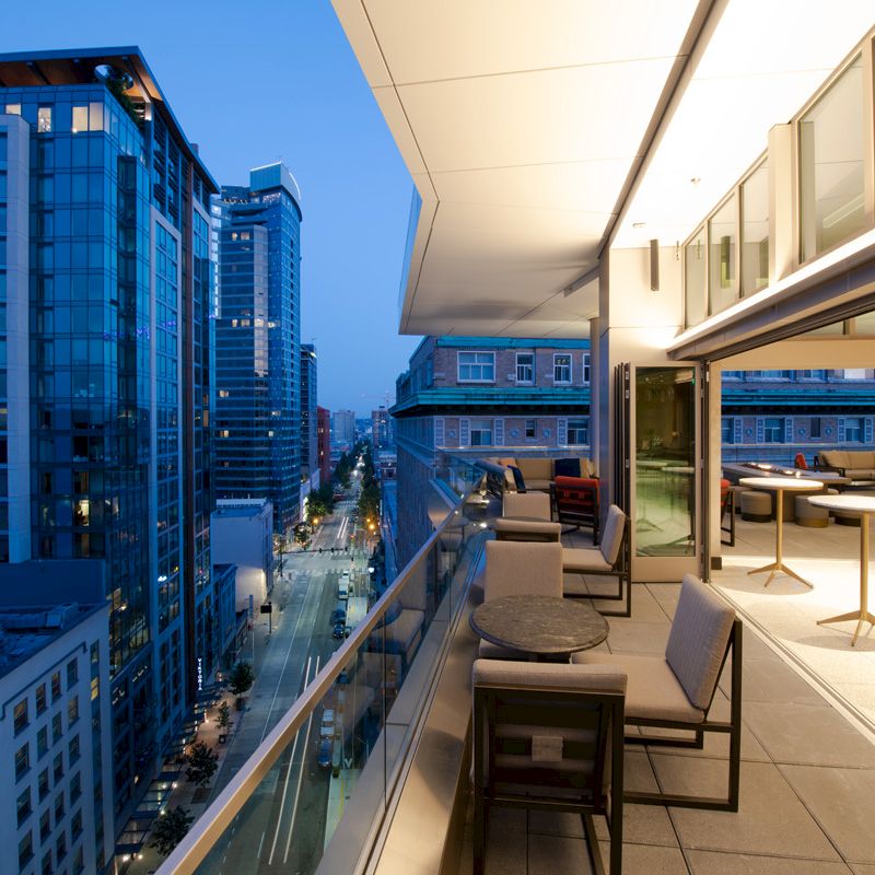 A modern rooftop terrace at dusk overlooking a cityscape with tall buildings. The terrace features seating areas and tables along the glass railing.