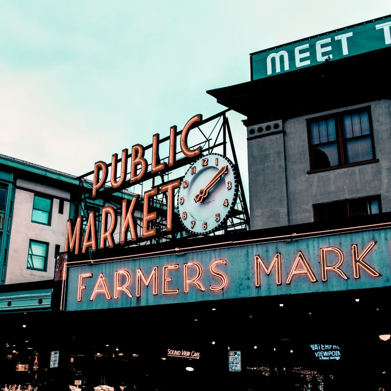 This image shows a public market with prominent neon signs reading "Public Market" and "Farmers Market" on a building facade.