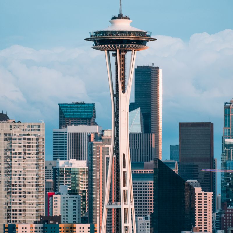 The image shows the Space Needle tower surrounded by various skyscrapers in the city of Seattle, Washington, under a partly cloudy sky.