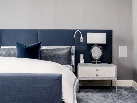 A modern bedroom with a navy blue upholstered bed, white bedding, a matching nightstand, a lamp, and a decorative piece. The room has a minimalist design.