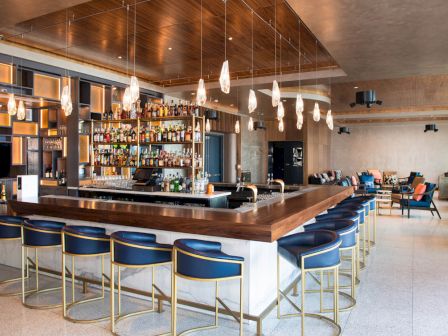 This image shows a modern bar with blue barstools, hanging pendant lights, a fully stocked bar shelf, and comfortable seating areas in the background.