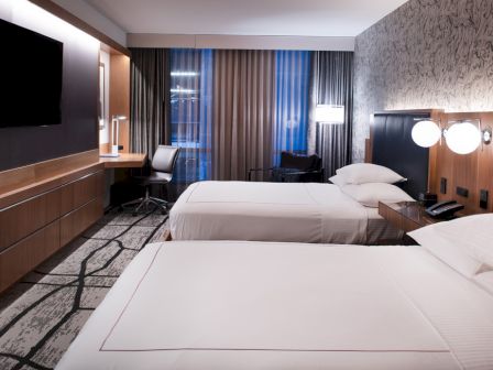This image shows a modern hotel room with two double beds, a desk with a chair, a TV on the wall, and stylish decor.