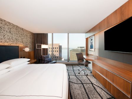 A modern hotel room features a large bed, wall-mounted TV, desk with chair, and a view of buildings outside through a large window.