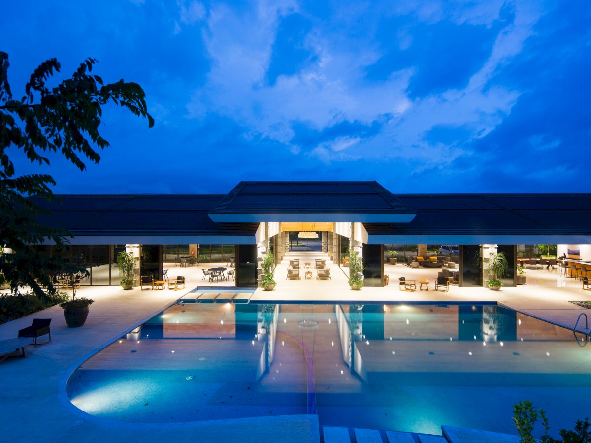 An illuminated pool area at dusk, with lounge chairs and a large building in the background, sky displaying hues of blue and purple at the day's end.