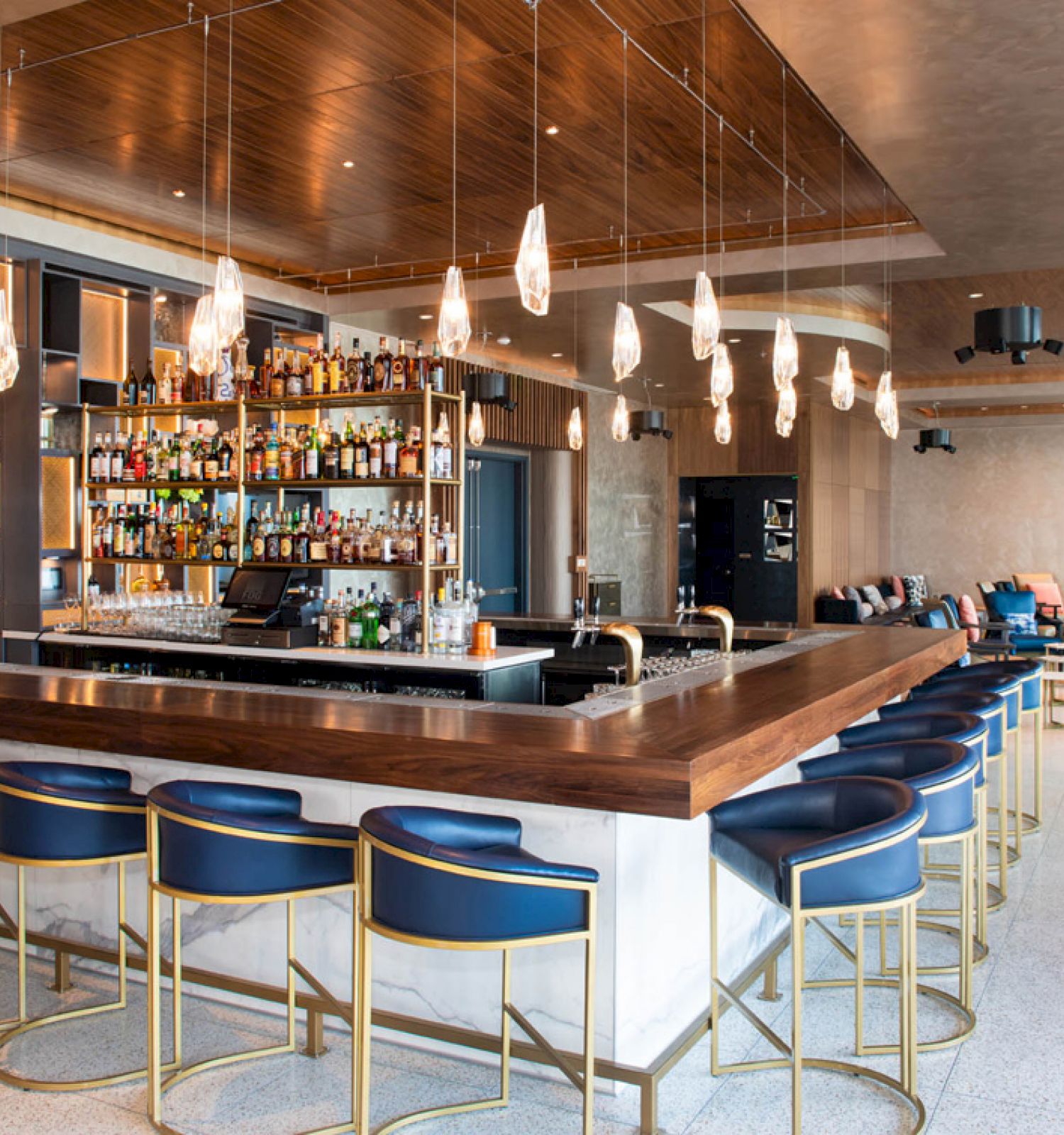 This image shows a modern bar with a wooden counter, blue-and-gold bar stools, and a well-stocked bar shelf, surrounded by comfortable seating.