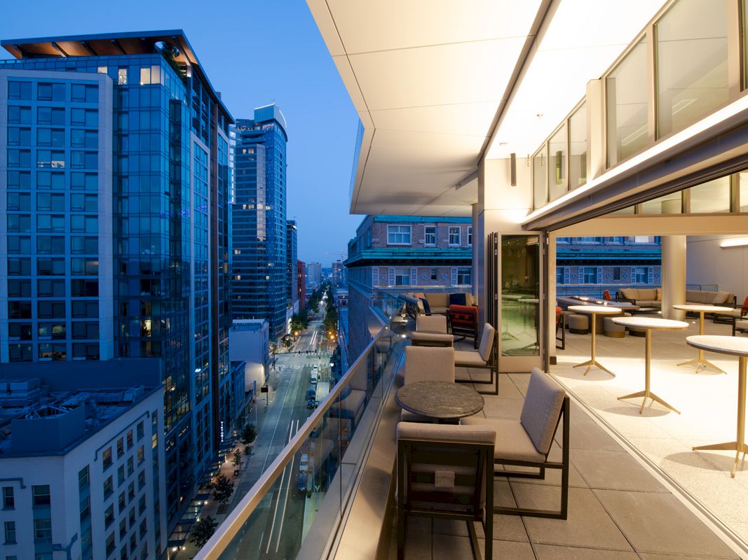 The image shows a modern cityscape at dusk, viewed from a high balcony with tables and chairs, overlooking a street lined with tall buildings.