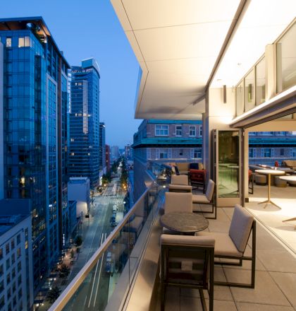 This image shows a modern cityscape at dusk, viewed from a high-rise balcony with outdoor seating and tables, overlooking a street with tall buildings.