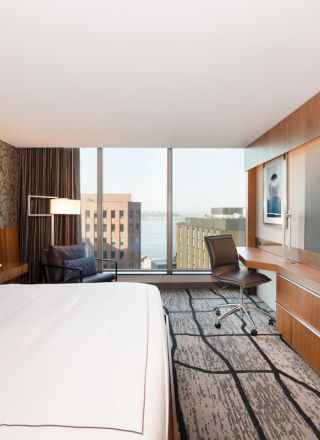 A modern hotel room features a large bed, a desk with a chair, a lounge chair, a TV, and a window offering a view of buildings outside.