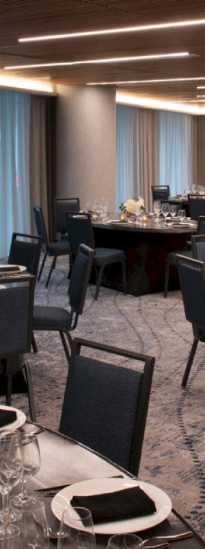 A banquet hall is set up with round tables, black chairs, plates, glasses, and silverware, decorated with white flowers and candles, under modern lighting.
