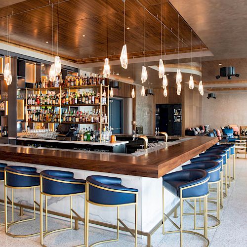 This image shows a modern bar with blue stools, a wooden counter, hanging lights, and a fully stocked bar. There is seating in the background.