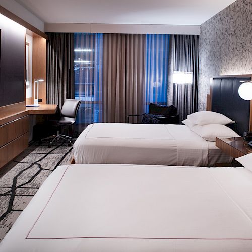A modern hotel room with two beds, a TV, a desk with a chair, and a well-lit window. The decor is contemporary with stylish lighting and furnishings.