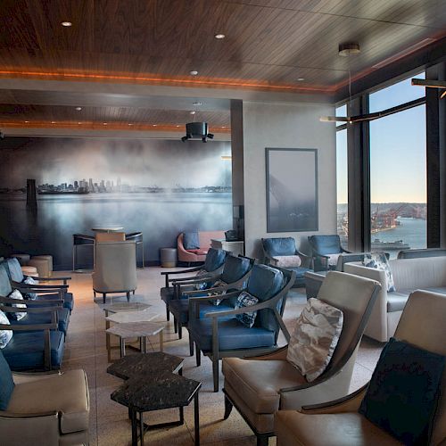 A modern lounge with leather chairs, a wall mural, and large windows offering views of a waterfront with a Ferris wheel, ending the sentence.