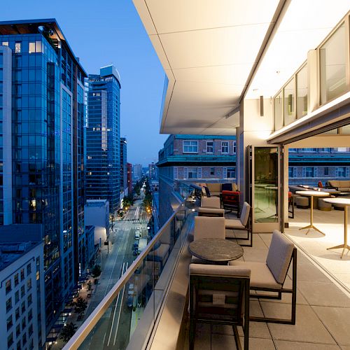 A modern balcony with chairs and tables overlooks a cityscape of tall buildings at dusk.