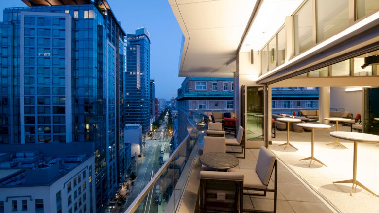A modern rooftop terrace with tables and chairs overlooks a cityscape with high-rise buildings and a street below.