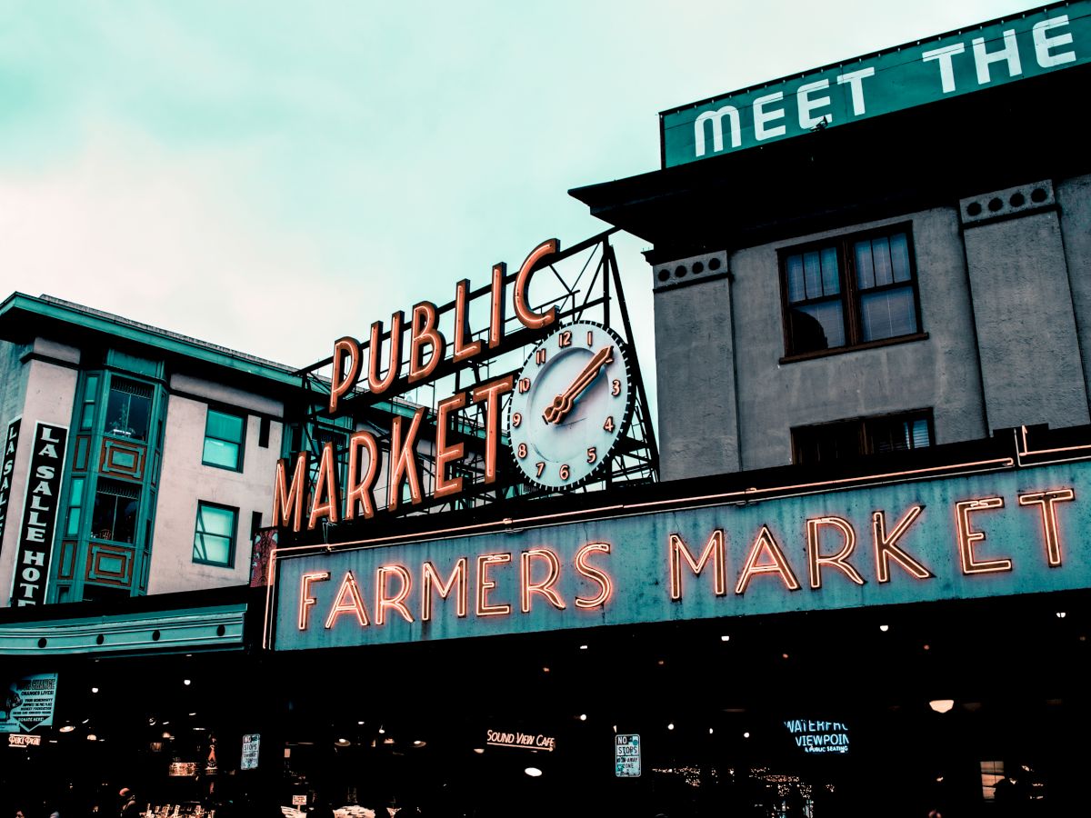 The image features a public market with neon signage that reads "Public Market" and "Farmers Market" against an urban backdrop of buildings.