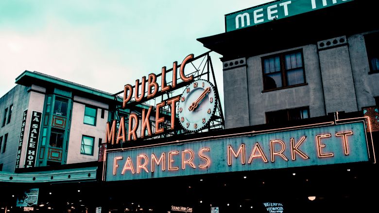 The image features a public market with neon signage that reads "Public Market" and "Farmers Market" against an urban backdrop of buildings.